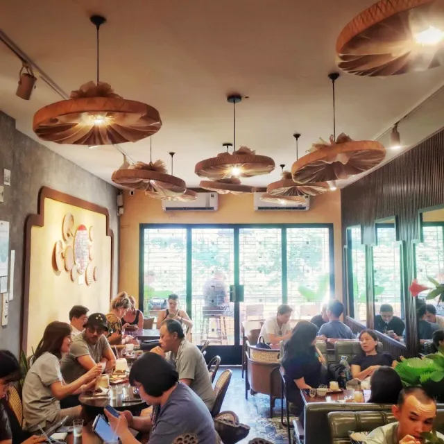 A busy space with large flat straw lampshades, guests around tables and glass doors to the terrace