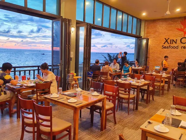A hall with glass doors open to the sea at sunset, wooden tables and a brick wall